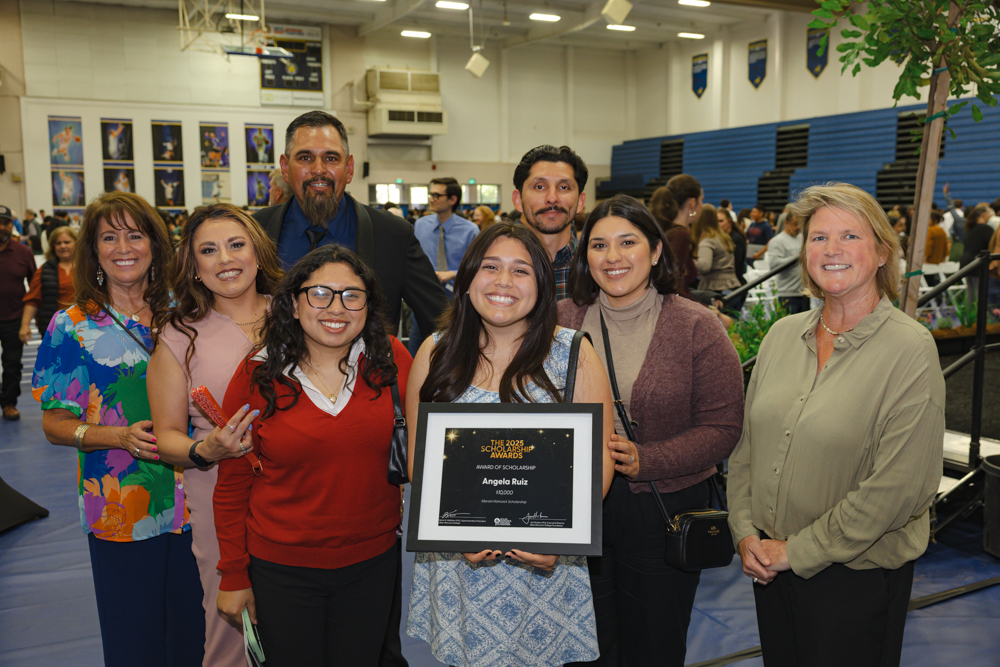A smiling student at Allan Hancock College’s 2025 Scholarship Awards Ceremony holds a framed certificate while surrounded by friends, family, and supporters inside a gymnasium. The group stands in front of a crowd of attendees and rows of blue bleachers.